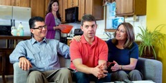 A husband sits uncomfortably between his in-laws while his wife notices from behind