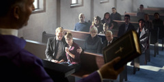 A priest holding a Bible as he conducts a sermon before mourners in a church.