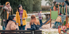 Four women of different racial backgrounds talking and laughing as their children play together at a playground.