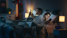 Mąż i żona leżą w łóżku późną nocą i każde z nich przegląda coś na swoim smartfonie.