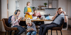 Uma mãe colocando a mesa do café da manhã e olhando irritada enquanto o marido e os filhos estão se distraindo com seus dispositivos eletrônicos.