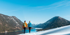 A couple walking on a snow-covered slope and looking at a lake surrounded by forested mountains under a clear blue sky.