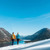 Семейна двойка се разхожда по заснежен склон и се любува на езеро, заобиколено от покрити с гори планини под ясно синьо небе.