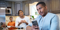 An annoyed husband using his smartphone while his wife tries to talk to him. Their young son is observing them.