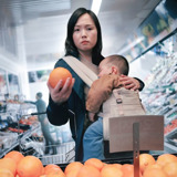 En un supermercado, una madre que lleva a su bebé dormido mira preocupada el precio de la fruta.