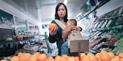 A mother anxiously looking at the price of some fruit while she grocery shops with her sleeping baby.