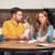 Parents discussing their finances at their dining table while their daughter is in the kitchen nearby.