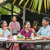 A family gathered around a table outside their modest home, happily sharing a meal with a guest.