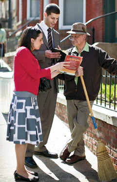 A couple, using the Bible and Bible-based publications, preaching to a man standing in front of his house.
