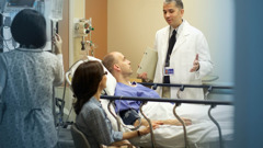 One of the doctors shown earlier, speaking with the patient and his wife in a hospital room.