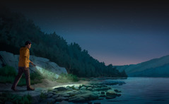 À la tombée de la nuit, un homme marche sur un chemin rocailleux le long d’un lac ; il éclaire devant lui avec une lampe torche.