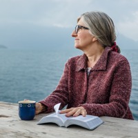A woman holds an open Bible, sits at a table, and stares into the distance