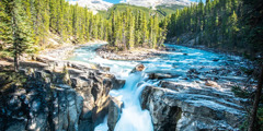 A river flowing toward a waterfall. The river is surrounded by snowcapped mountains and a forest.