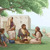 Una familia israelita sentada bajo un árbol afuera de su casa conversando alegremente. Unas ovejas pastan cerca.