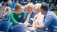 Nuori pariskunta juttelee iloisesti iäkkäämmän pariskunnan kanssa aluekonventissa.