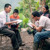 A brother reading a scripture to a family outside their modest home. The father follows along in his Bible while the mother and young son listen intently.