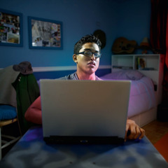 A young man, alone in his bedroom, looking away from his computer to avoid viewing something that popped up on the screen.
