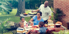 A happy family eating together outdoors. The father grills and puts food on a table.