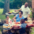 Eine glückliche Familie sitzt gemeinsam beim Essen im Garten. Der Vater grillt und bringt das Essen zum Tisch.