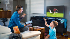 Un hermano comiendo y viendo la televisión mientras hace su estudio personal. Su hijo está a su lado jugando.