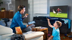 Un germà menjant i mirant la televisió mentre fa estudi personal. El seu fill està jugant al seu costat.