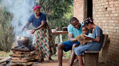 A family being content outside their modest home. The mother cooks a simple meal over a fire. Nearby, the father uses the book “Lessons You Can Learn From the Bible” to study with their two children.