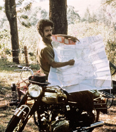 Робърт стои до мотоциклета си и показва на карта къде е пътувал.