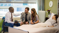 Старейшина утешава семейна двойка с помощта на Библията. До тях лежи момиче на болнично легло.