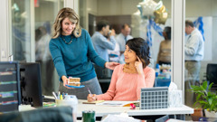 A workmate offering a sister a piece of birthday cake at her desk. Others eat, drink, and celebrate in a neighboring office.