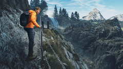 Den unge mannen på bildene i de forrige artiklene går på en smal sti langs en fjellskrent og holder seg i et kjettinggjerde. I bakgrunnen ser man en hengebro og en fjelltopp.