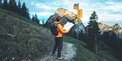 Un home jove mirant un mapa mentres està parat en una senda de muntanya. En front d’ell, té un pal amb molts senyals que apunten en direccions diferents i en la distància, es pot vore el cim d’una muntanya.
