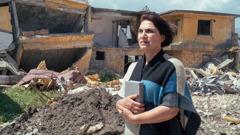 Una hermana de pie al lado de una casa que fue destruida por un desastre sostiene una Biblia y demuestra confianza.