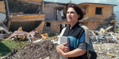 Сестра стоит рядом с домом, который был разрушен стихийным бедствием, и держит в руках Библию. На её лице спокойствие.