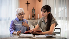 A sister conducting a Bible study with an older woman who has a cross on the wall and an idol on a table. The woman is amazed to see Jehovah’s name in her own Bible.