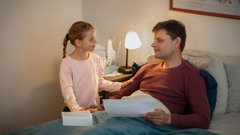 A brother in bed holding a sheet of paper as his young daughter holds his hand.