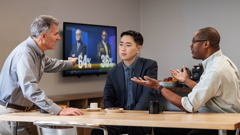 A brother taking a break with two coworkers who are forcefully expressing their opinions. One of the coworkers gestures toward a TV that shows the poll results for two political candidates.