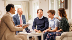 Dos ancianos reunidos con un menor bautizado y con sus padres en la casa de ellos. Uno de los ancianos les está leyendo un texto de la Biblia.