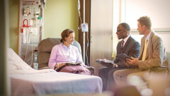 Dois anciãos visitando uma irmã no hospital. Eles estão com suas Bíblias abertas conversando com a irmã.