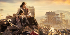 A woman with her arm around her young daughter while they sit on a pile of rubble in a city that has been devastated by war. They are surrounded by destroyed buildings.