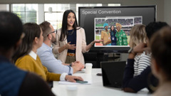 A reenactment of Angelena presenting a slideshow to her coworkers. She happily shows a photo of herself at a special convention, posing with delegates from another country.