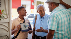Молодой брат проповедует по домам вместе с пожилым. Пожилой брат разговаривает с мужчиной, а молодой брат стоит рядом, и у него на планшете открыто видео.