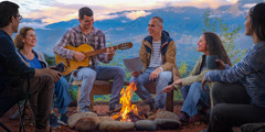 Amigos de diferentes idades sentados à volta de uma fogueira. Um deles está a tocar guitarra.