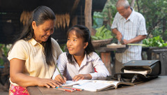 A mother helping her young daughter with her homework while the father cuts up vegetables nearby.