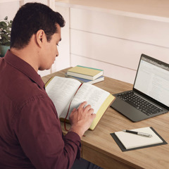一名男子一边看圣经一边用电脑搜集资料。在他手边有笔和纸以及其他书本。