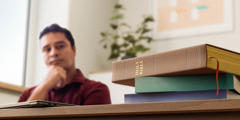 Мужчина задумчиво смотрит на Библию и другие книги, которые лежат на столе.
