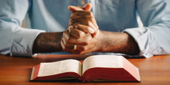 A man with clasped hands, praying. His Bible is open on the table in front of him.