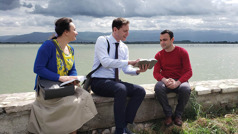 Alida and Stefano preaching to a man as they all sit on a stone wall by a lake.