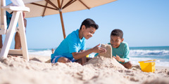 Children building a sandcastle at the beach as their father sits nearby.