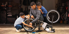 A father showing his two young sons how to repair a bicycle.
