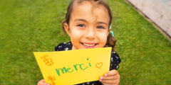 A young girl, with a big smile on her face, holding up her hand-drawn thank-you note.